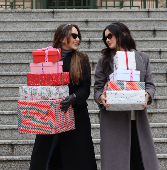 Două femei elegante țin în brațe cutii cadou frumos ambalate, pregătite pentru sezonul de sărbători – inspirație pentru idei de cadouri beauty de la WowSkin